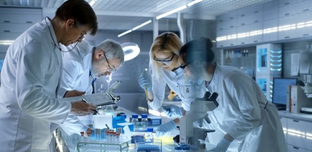 A group of scientists in white lab coats lean over a sample in a lab.