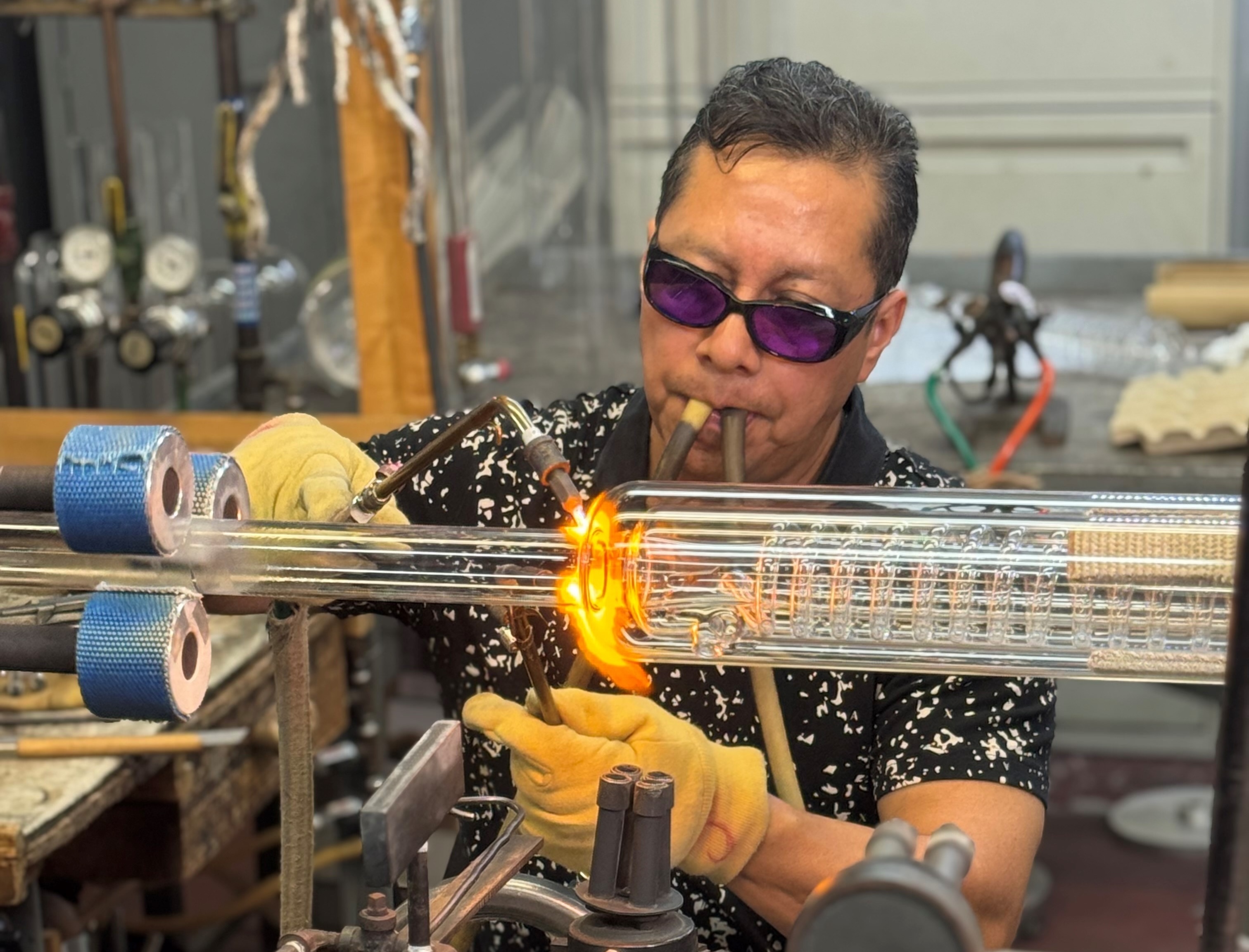 glass blowing apparatus tool blow torch Wilmad A man heating a glass apparatus with a blow torch. He has goggles on and two tubes in his mouth.