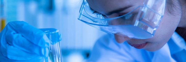 A scientist wearing clear lab goggles looking at a vial of solution.