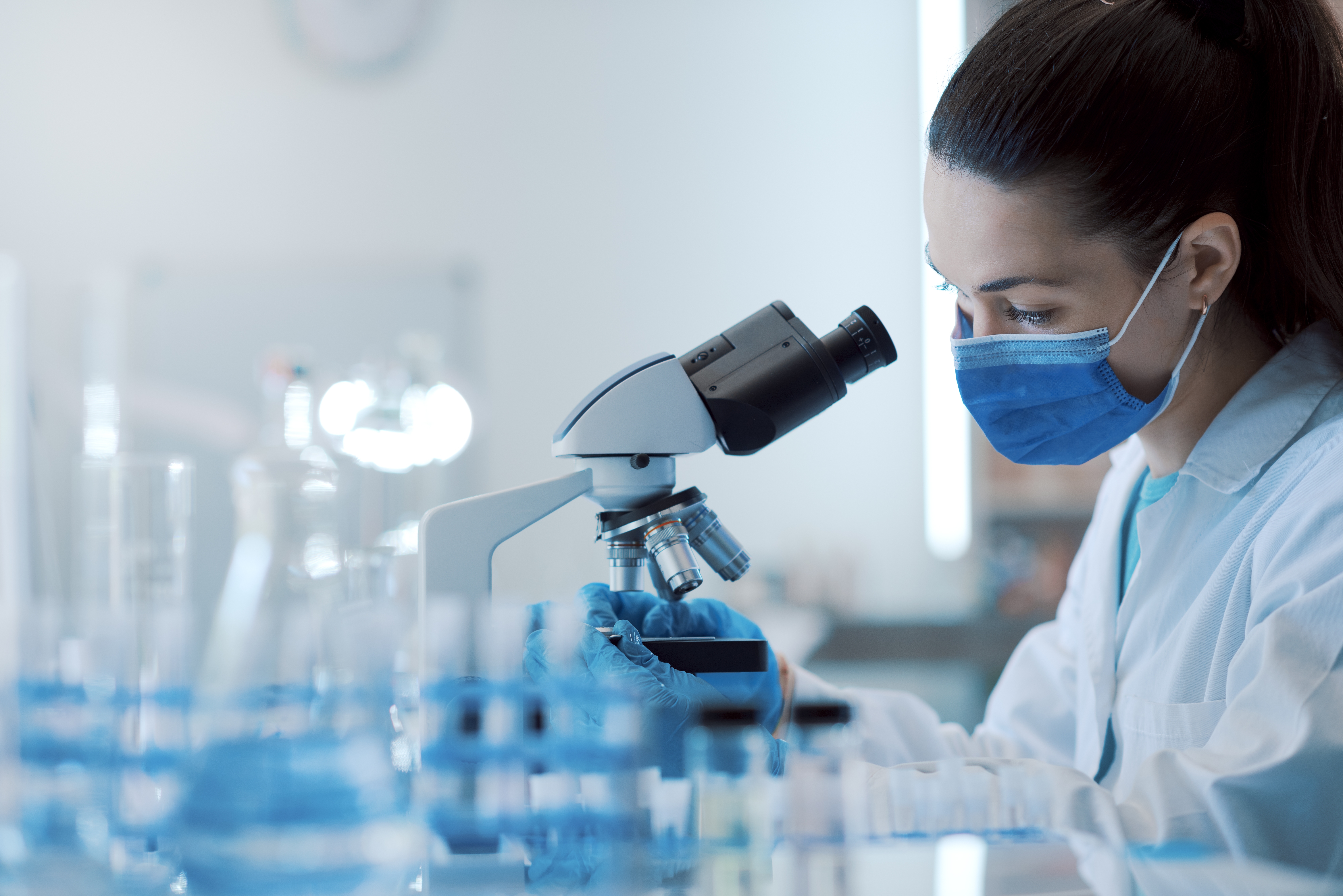 A scientist in a lab wearing a mask looking through a microscope.