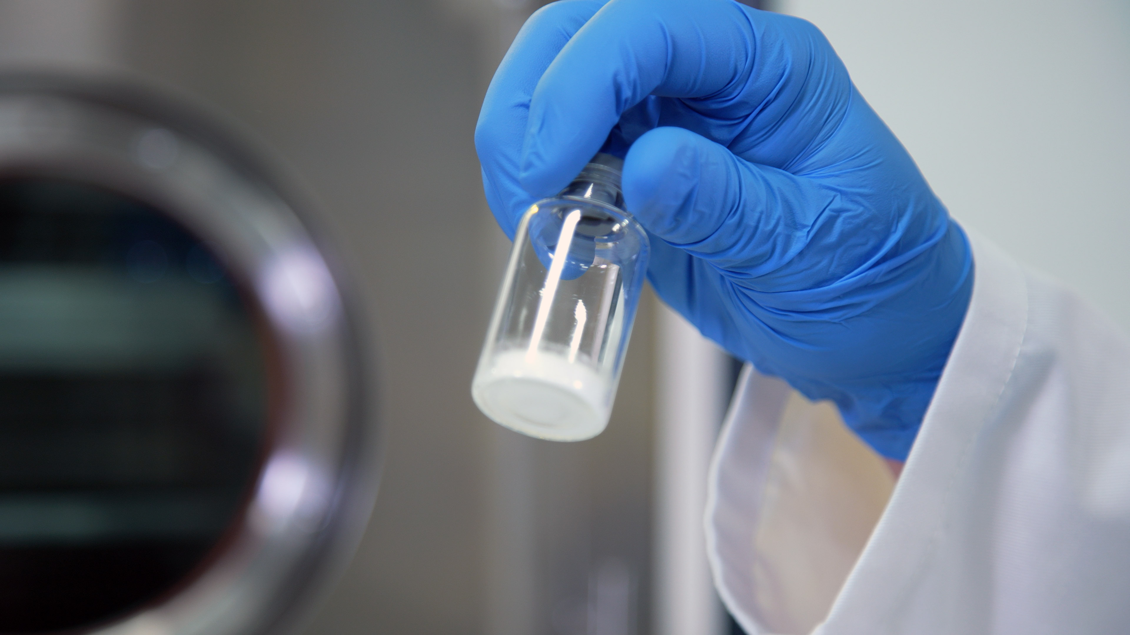 A gloved hand holding a clear vial with a bit of white powder at the bottom.