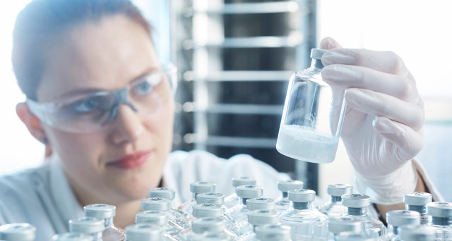 A scientist with goggles on examines the product of a freeze-drying cycle in a large glass vial.